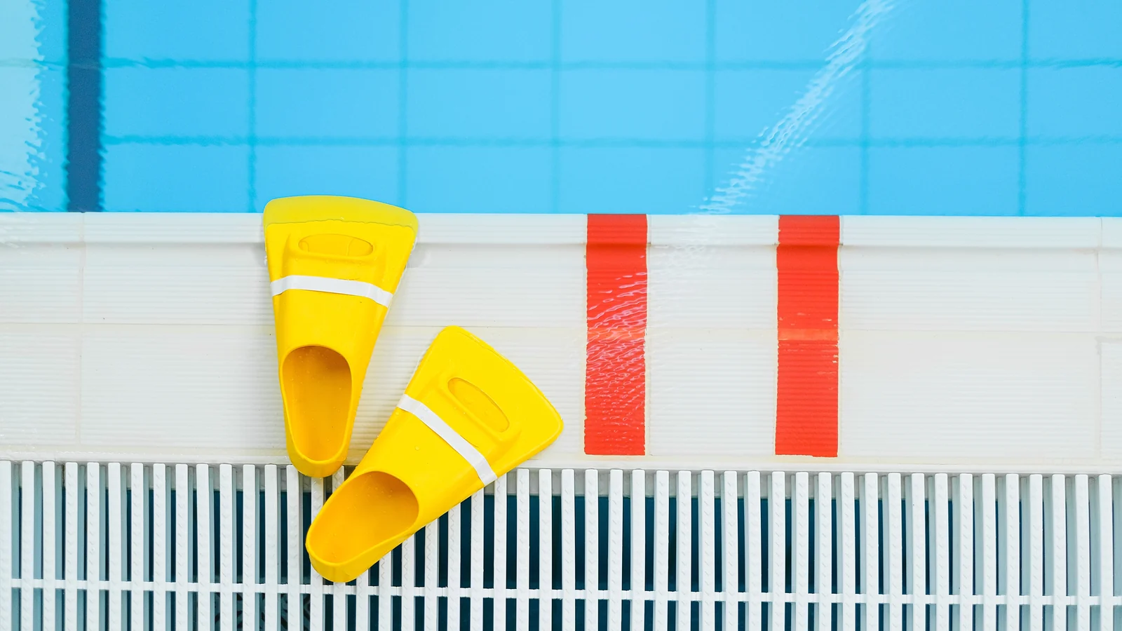 Gelbe Schwimmflossen auf einem weißen Beckenrand neben blauem Poolwasser und roten Markierungen.